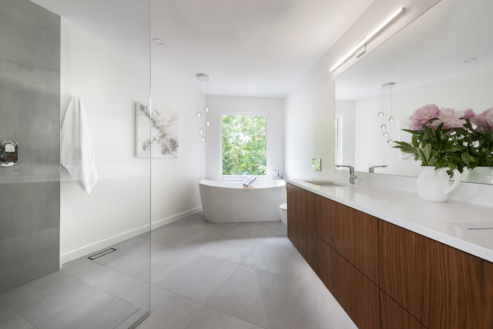 Modern Scandinavian bathroom with a freestanding white tub, large window, floating walnut wood vanity with dual sinks, and a curbless glass shower. The space is bright and minimalist, featuring clean lines, light gray tiles, and elegant pendant lighting.