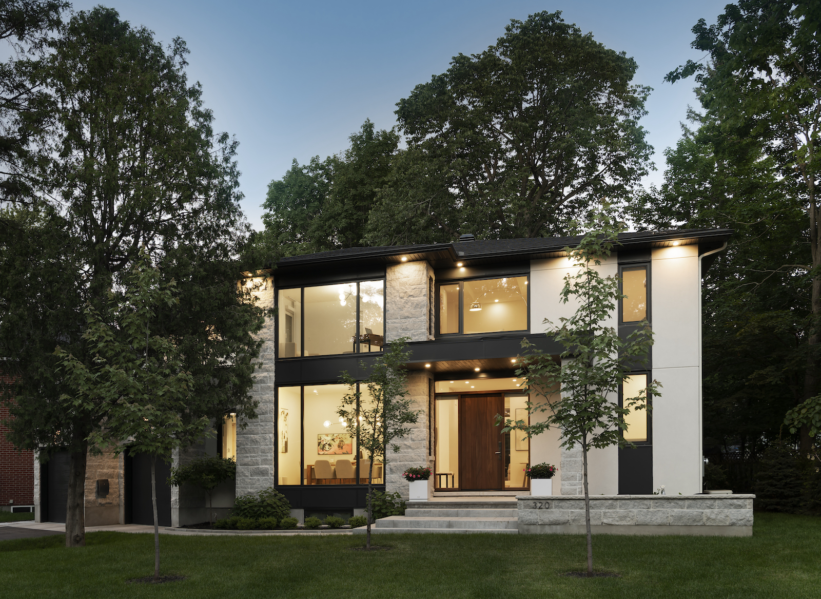 Modern Scandinavian-style two-story home with large windows, light stone exterior, and black accents. The house features a welcoming entrance with a wooden door, potted plants, and exterior lighting. Surrounded by mature trees, the design emphasizes natural light and a connection to the surrounding greenery.