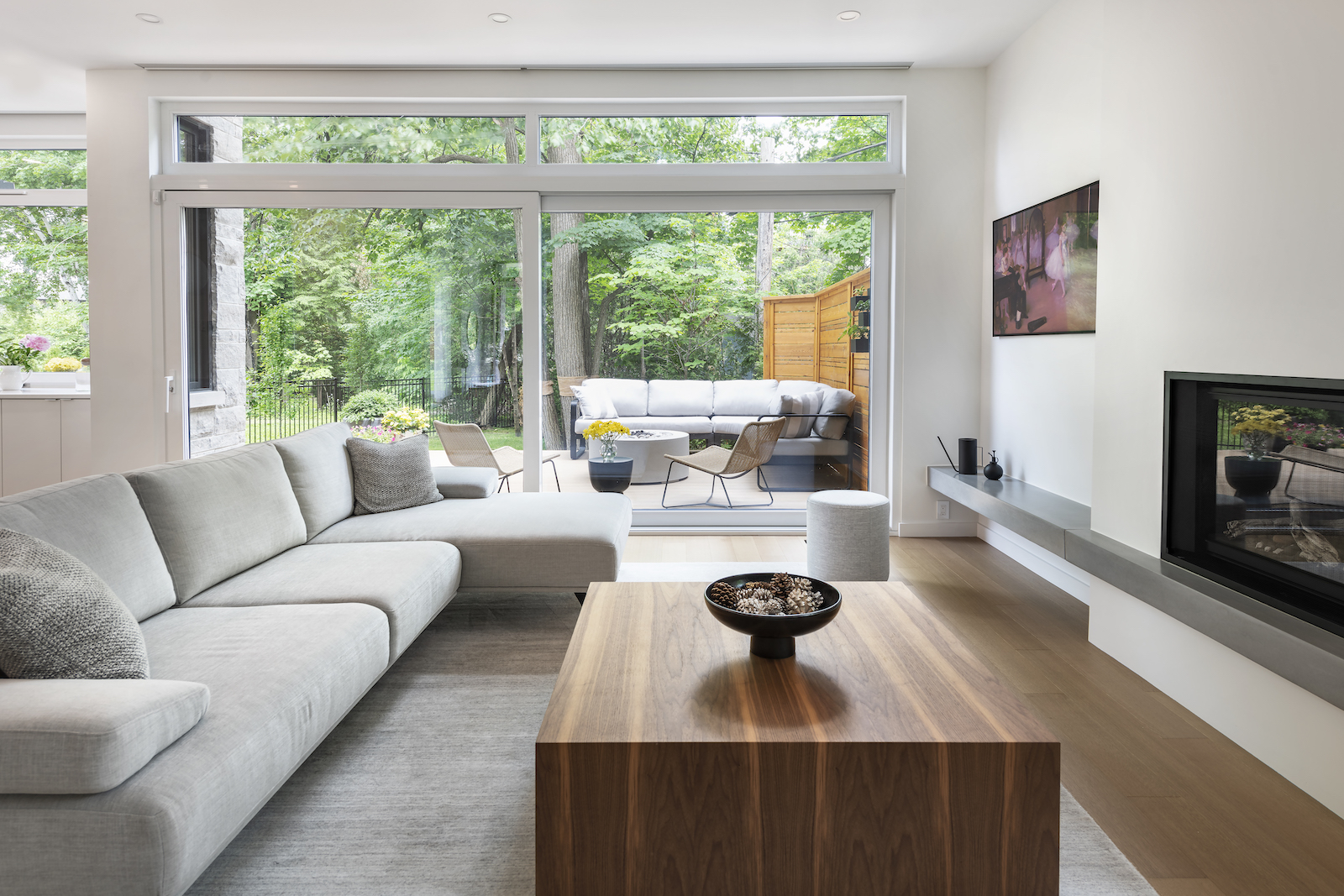 Modern Scandinavian living room with a large gray sectional sofa, modern walnut coffee table, built-in fireplace, and floor-to-ceiling windows overlooking a green backyard with outdoor seating.