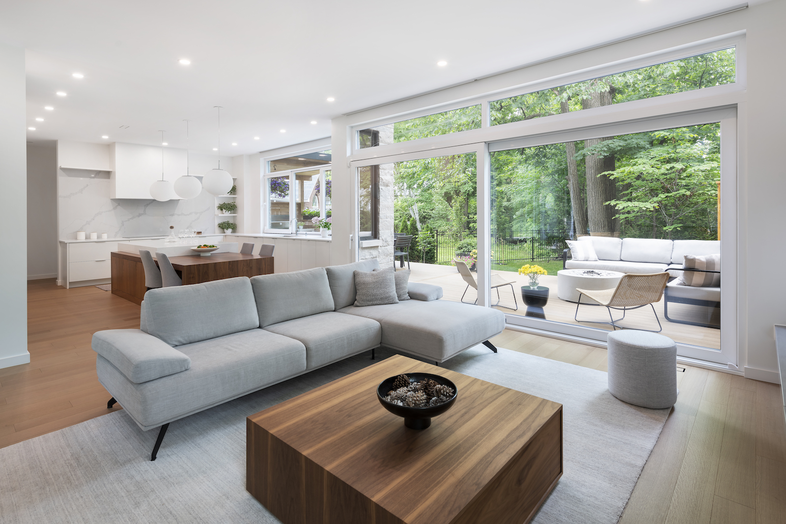 Modern Scandinavian open-concept living space with a large gray sectional sofa, modern walnut coffee table, and a seamless transition to the kitchen area. The room features floor-to-ceiling windows that open to a green backyard with outdoor seating, blending indoor and outdoor living