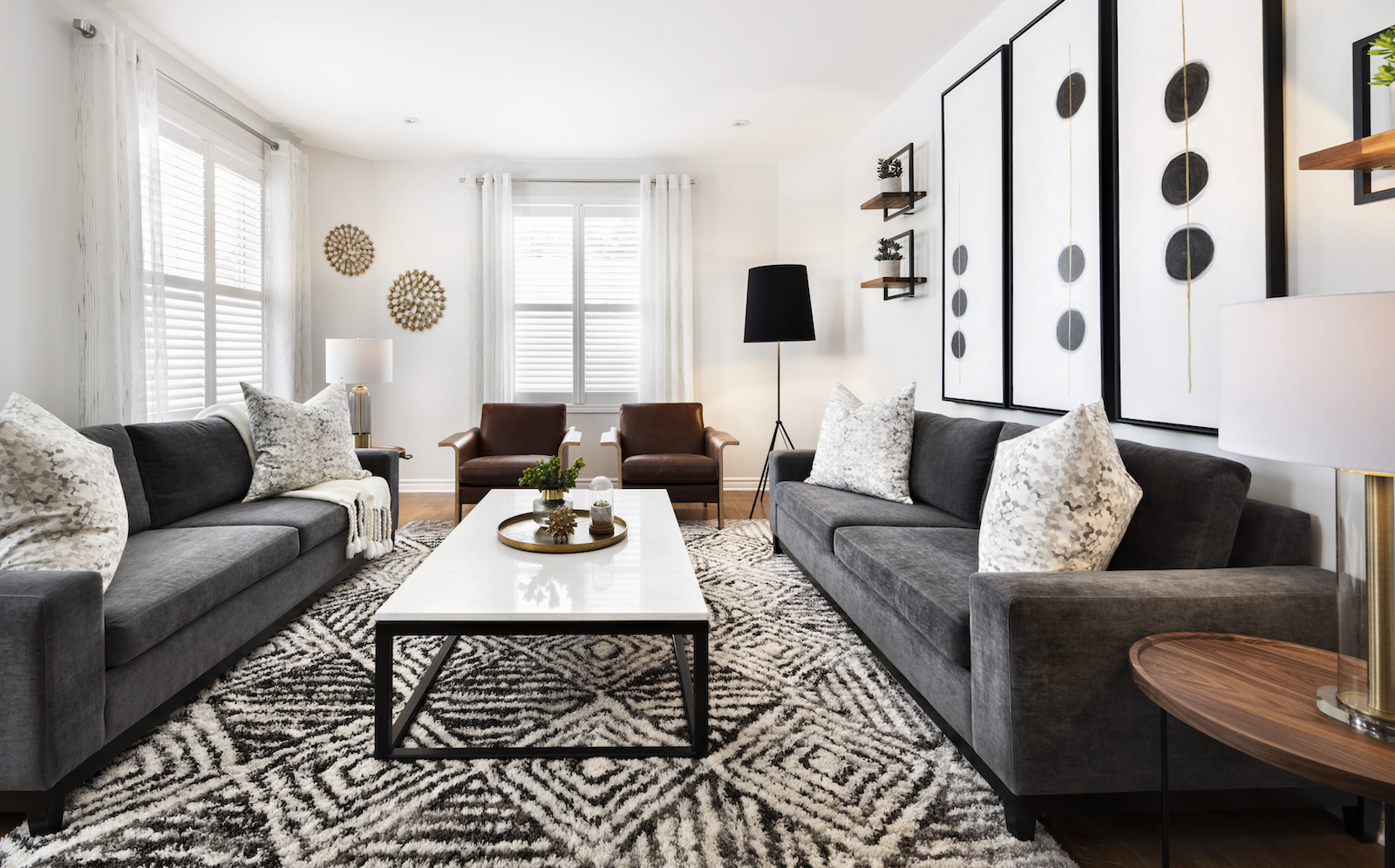 Modern living room in Ottawa designed by an interior decorator, featuring mid-century modern style with charcoal grey sofas, saddle tan leather chairs, large black and white geometric wall art, white coffee table, patterned rug, and neutral tones throughout.