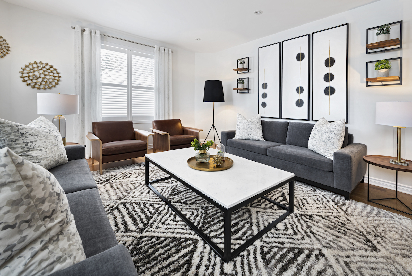 Modern living room in Ottawa designed by an interior decorator, featuring mid-century modern style with charcoal grey sofas, saddle tan leather chairs, large black and white geometric wall art, white coffee table, patterned rug, and neutral tones throughout.