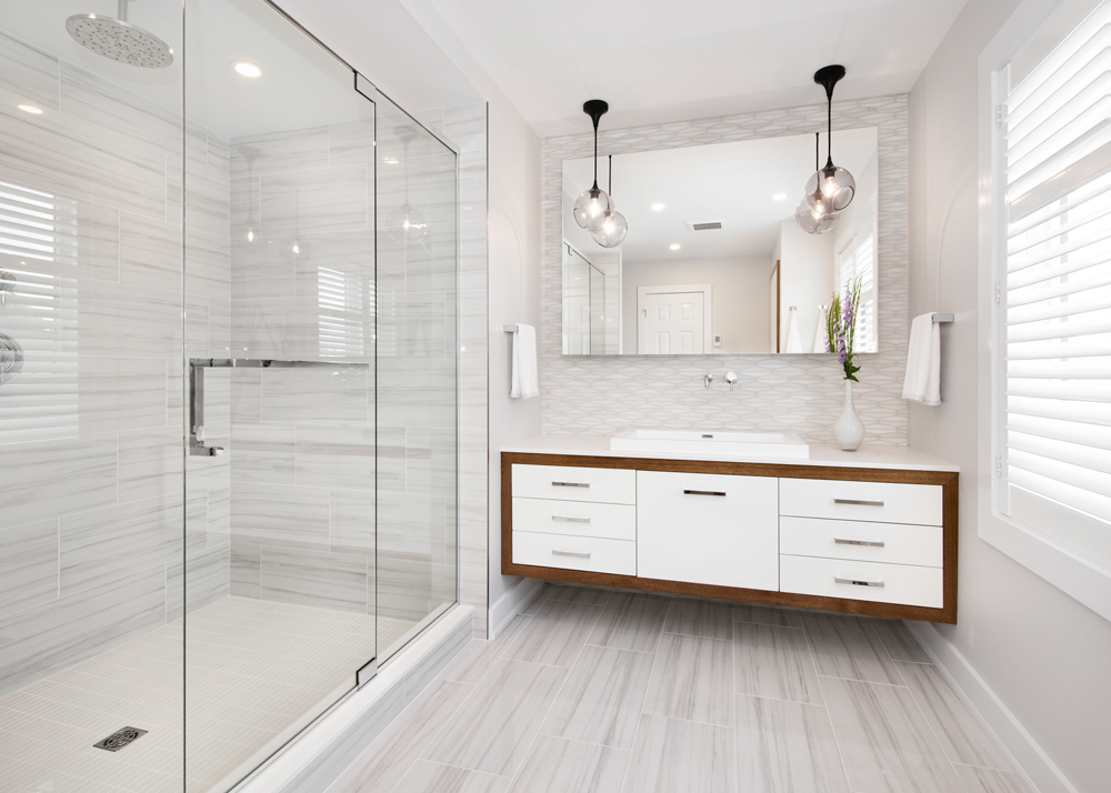 Modern bathroom in Ottawa designed by a bathroom designer and interior designer, featuring a large glass-enclosed shower, custom floating vanity with a two-tone finish, wall-mounted faucet, floating mirror, and chrome fixtures. The space includes soft grey tile flooring and pendant lights for a sleek, spa-like atmosphere.