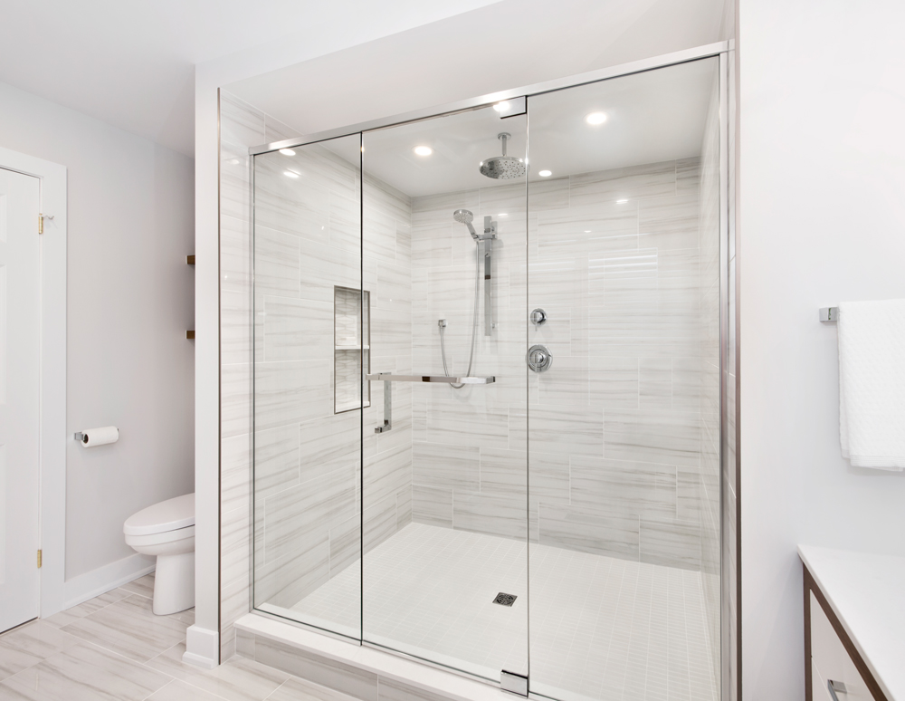 Modern bathroom in Ottawa designed by a bathroom designer and interior designer, featuring a large glass-enclosed walk-in shower, marble-look tiles, chrome fixtures, and recessed lighting. The design showcases a sleek, minimalist aesthetic with soft grey tones and a built-in shower niche for added convenience.