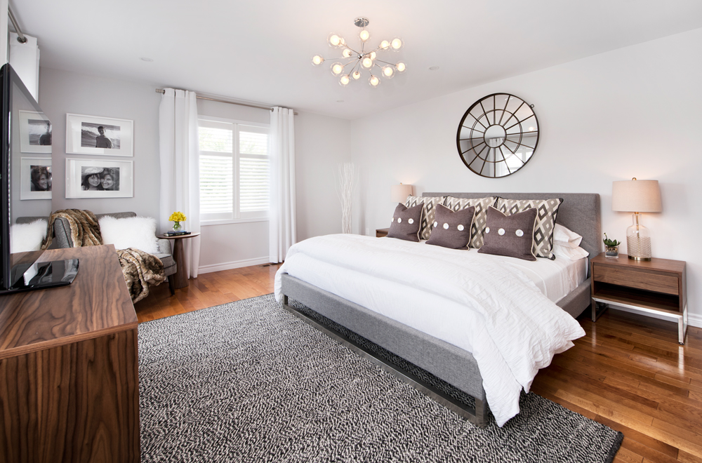 Modern bedroom designed by an interior decorator in Ottawa, featuring a grey upholstered bed, starburst chandelier, round mirror, wooden nightstands, and a textured grey rug with mid-century modern touches.