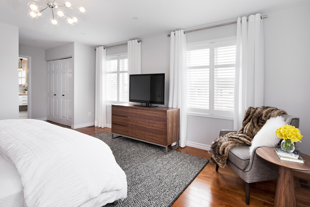 Modern bedroom designed by an interior decorator in Ottawa, featuring a grey upholstered bed, starburst chandelier, round mirror, wooden nightstands, and a textured grey rug with mid-century modern touches.