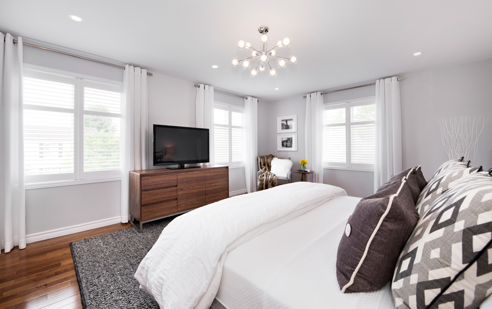 Modern bedroom designed by an interior decorator in Ottawa, featuring a grey upholstered bed, starburst chandelier, round mirror, wooden nightstands, and a textured grey rug with mid-century modern touches.