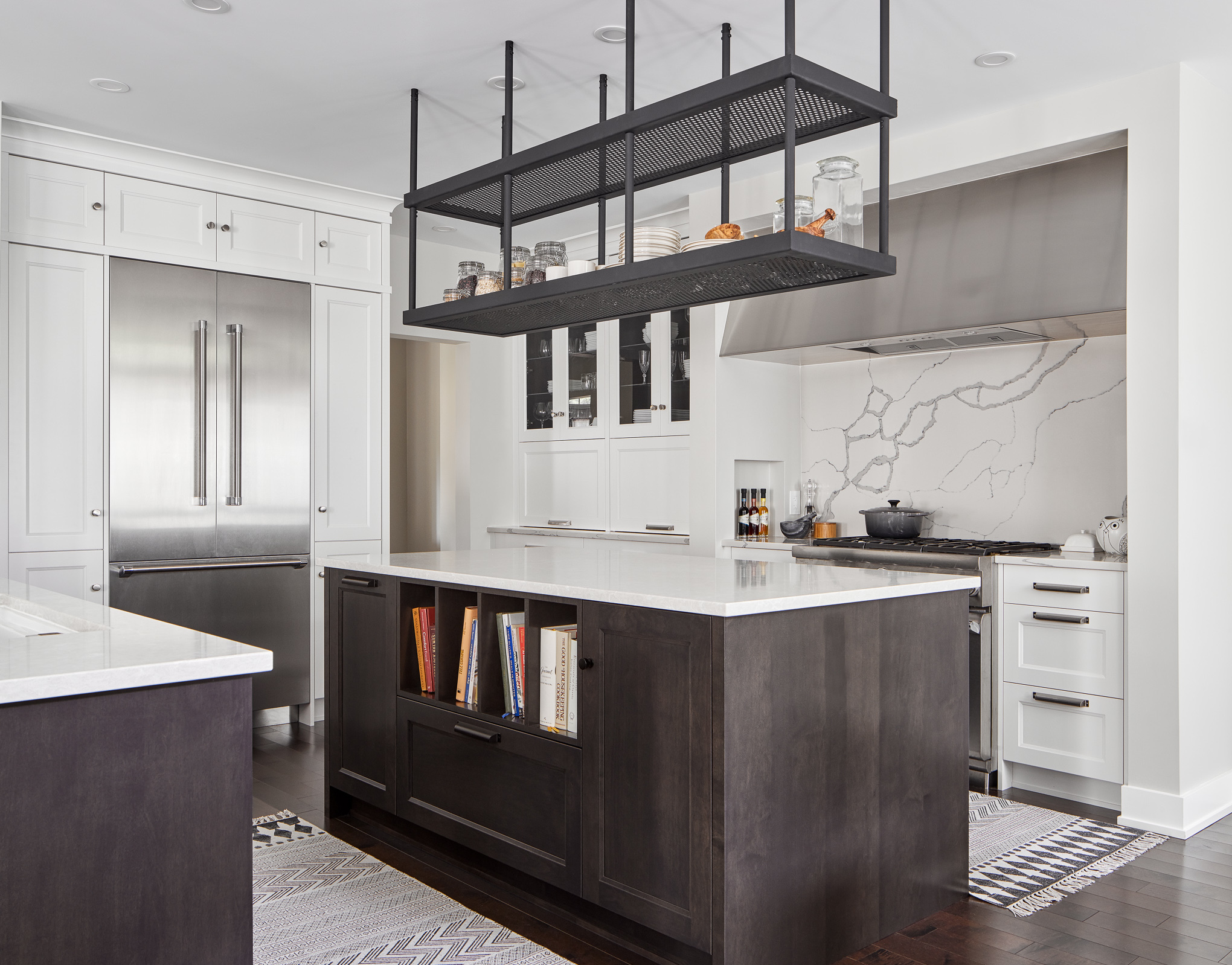 Transitional kitchen with dual islands, custom stainless steel hood fan, veined quartz backsplash, and industrial hanging shelving. Features dark wood island, built-in refrigerator, and custom cabinetry. Designed by Ottawa kitchen designers.