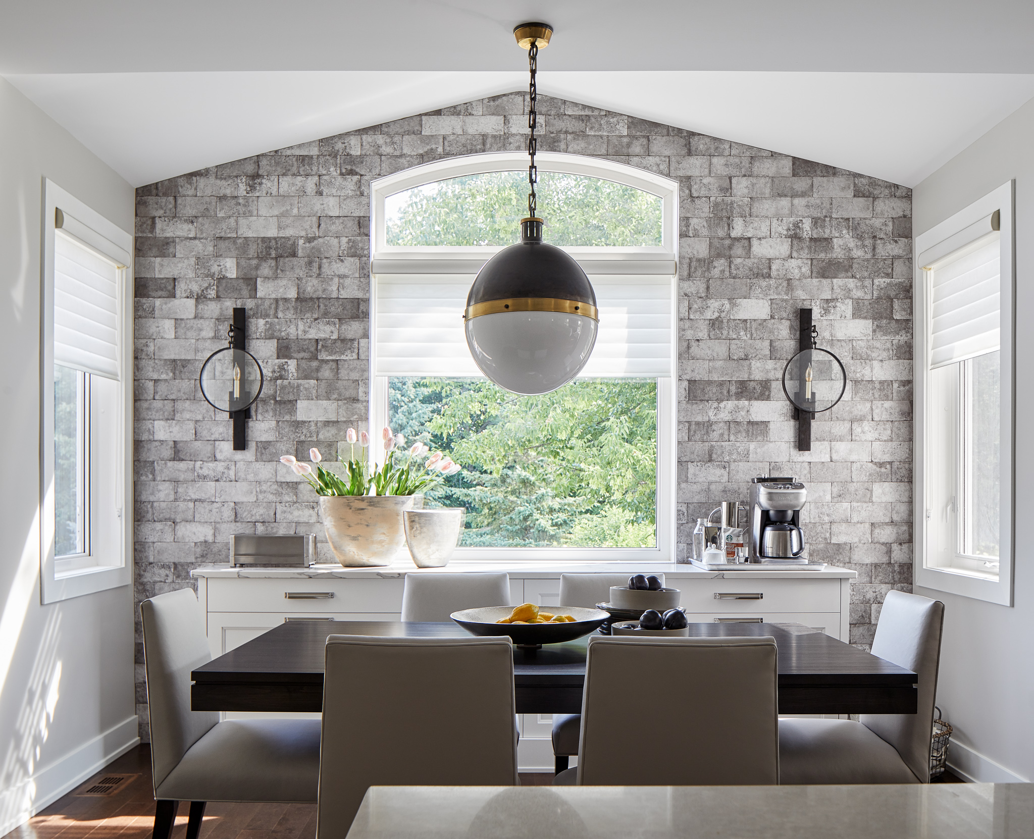 Dining area with faux stone accent wall, large window, and modern pendant light. Features a dark wood dining table, light upholstered chairs, and natural light. Designed by Ottawa kitchen designers.