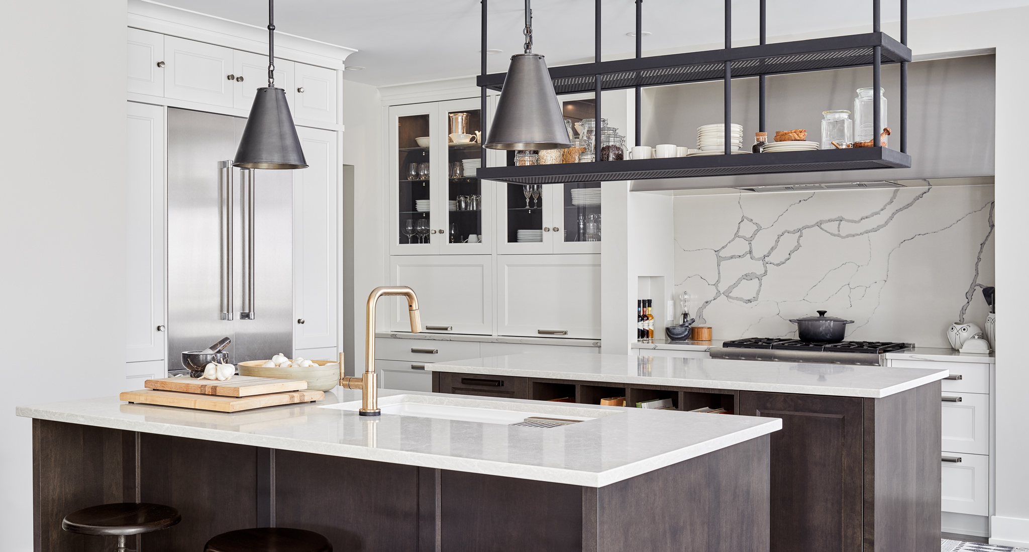 Transitional kitchen with dual islands, veined quartz backsplash, custom stainless steel hood fan, and industrial hanging shelving. Features custom cabinetry, built-in appliances, and a brushed brass faucet. Designed by Ottawa kitchen designers.